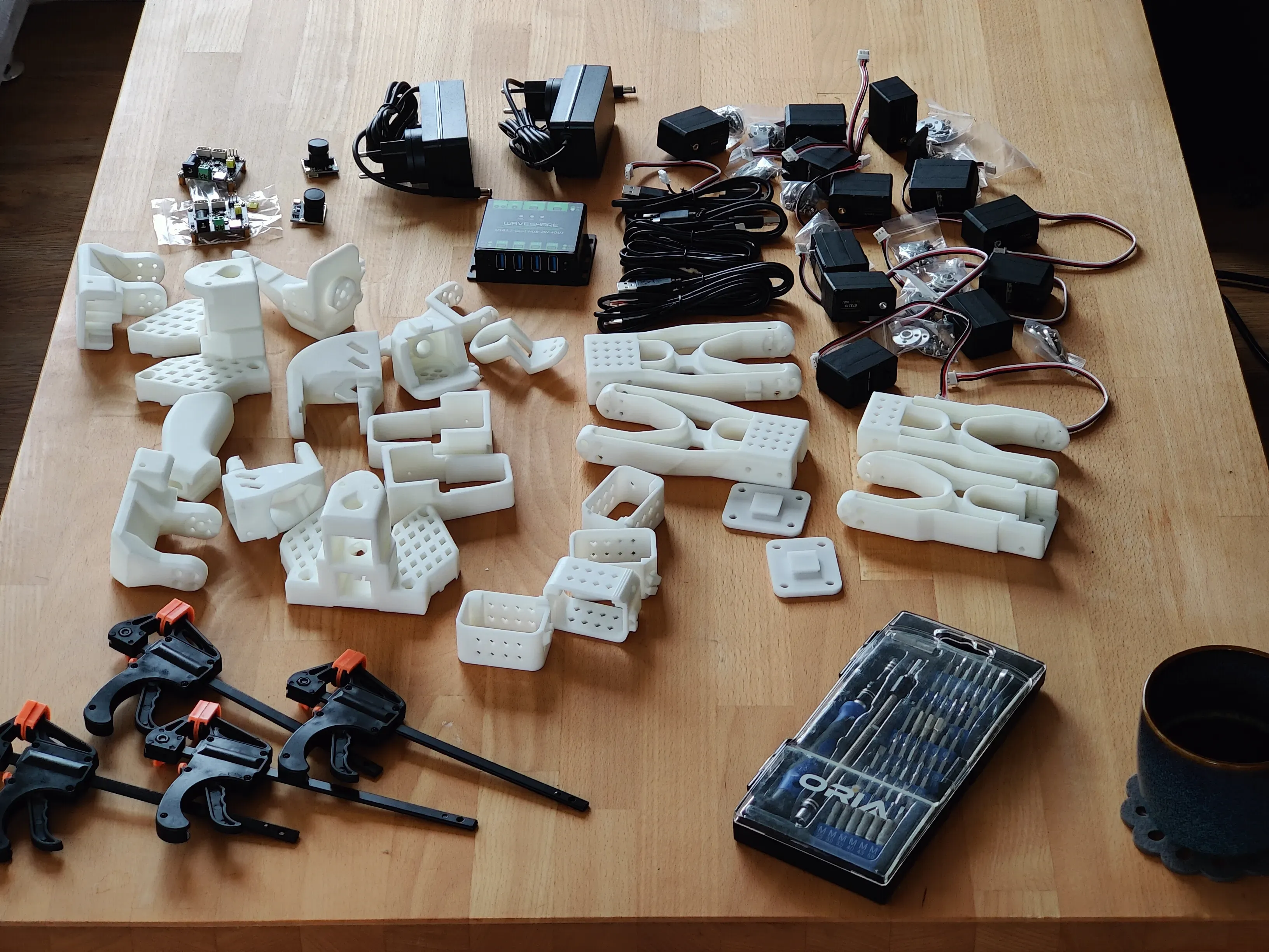 Printed arm parts, servos, power supplies, a USB hub, cameras, clamps and a screwdriver set laid out on a table